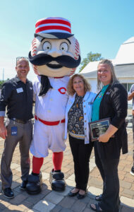 Three adults stand smiling outdoors with a baseball mascot dressed in a Cincinnati Reds uniform. One woman holds the Ability Transcends Challenges Award plaque from Project LIFE as everyone poses together on a sunny day.