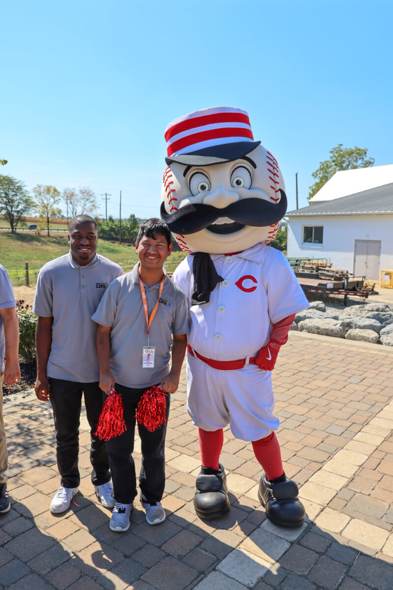 Three people stand outside on a stone patio. Two are smiling adults in gray shirts, one holding red pom-poms and a Project LIFE sign, celebrating the 2025 Ability Transcends Challenges Award. Beside them is a tall baseball mascot in a striped hat and white uniform with a red “C.”.