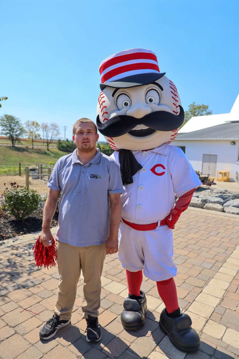 A man stands next to a person in a Cincinnati Reds mascot costume, celebrating the 2025 Ability Transcends Challenges Award. The man holds red pom-poms and wears beige pants and a gray polo on this sunny Project LIFE event day outdoors.