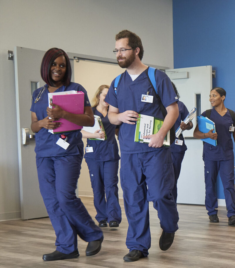 A group of nursing students in blue scrubs study, talk, and walk in a modern lounge with a blue wall displaying the Butler Tech logo. The welcoming space highlights adult education and offers student support as they read books or discuss coursework.