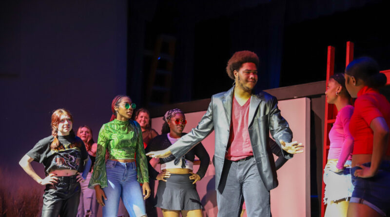 A group of young performers stand on stage, immersed in the performing arts. One person in a black jacket and red shirt speaks animatedly to others in colorful, casual outfits as they watch attentively under bright stage lighting.