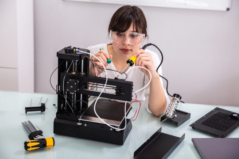 A person wearing safety goggles works on assembling a 3D printer at a desk, applying Electrical Technology skills with tools and a soldering iron. Various components and parts related to power systems are scattered across the glass surface.