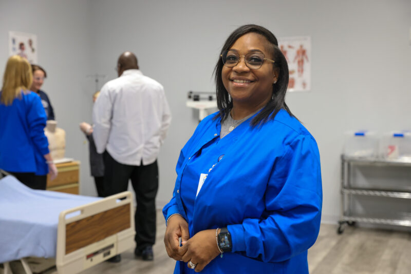 A woman in blue scrubs smiles at the camera in a medical training room, showcasing an Adult Education setting where others interact near a hospital bed and medical equipment in the background.