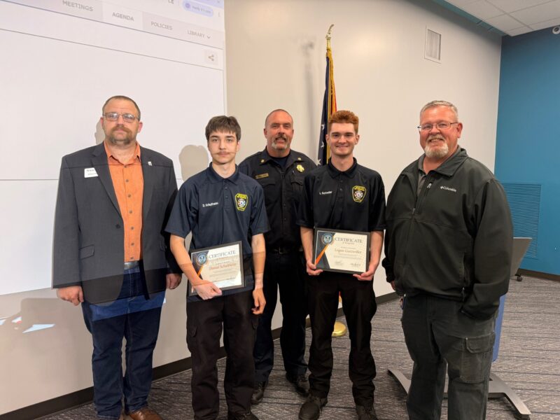 Five men stand indoors; two Butler Tech Students in the center hold certificates and wear matching uniforms with police badges. Behind them is a person in a police uniform. The group is smiling, with an American flag in the background.