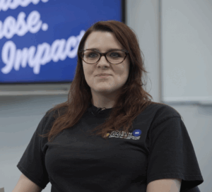 A woman with long brown hair and glasses sits at a desk, smiling. She wears a black shirt with a Butler Tech logo. Behind her, a blue screen displays the words "Passion. Purpose. Impact." in white text.