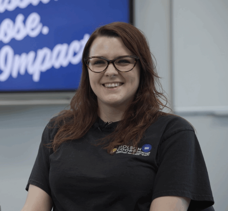 A smiling woman with long brown hair and glasses sits indoors, wearing a black Butler Tech t-shirt with a logo. Behind her, a blue screen displays the words 