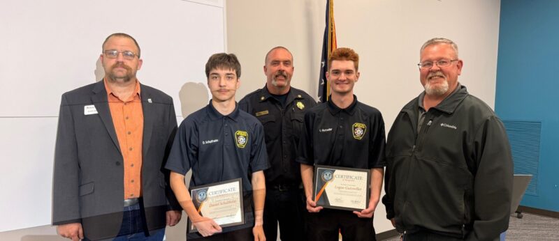 Five men stand together indoors, two of them holding certificates and wearing matching uniforms, while the others wear business or casual attire; a flag is in the background.