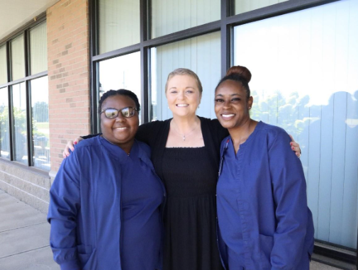 Three women stand outside a building, smiling at the camera. Two wear navy blue scrubs from Butler Tech, while one in a black dress has short, light-colored hair. United, they embody student care and support for one another.
