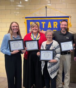 Four adults stand smiling indoors, each holding a certificate of appreciation. They are in front of a brick wall and a Rotary Club banner. The group consists of three women and one man, all dressed in business casual attire.