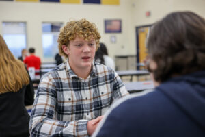 A teenage boy with curly blond hair wearing a plaid shirt sits across from another person at a cafeteria table, engaged in conversation that builds student confidence. Other people are visible in the background.