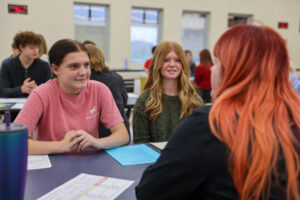 Three teenagers sit at a table having a conversation in a classroom or cafeteria setting. Two girls face the camera while a third, with bright red hair, sits with her back to the camera. They appear to be practicing mock interviews to build student confidence.