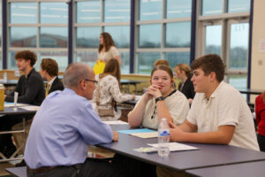 Students and adults sit at tables in a bright, modern room, practicing mock interviews and building career skills. Papers and water bottles are on the tables, sunlight pours through large windows, and a woman stands in the background.