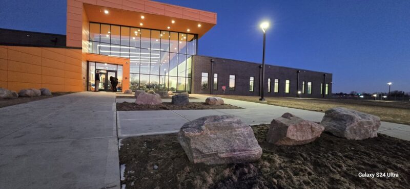A modern Aviation Center with large glass windows and orange accents is lit up at dusk. Several people are visible inside. The sidewalk and scattered large rocks lead to the entrance, where a streetlamp glows—perfect for aspiring flight training students.