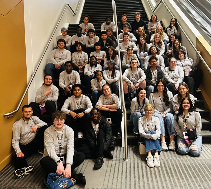 A large group of people wearing matching gray SkillsUSA shirts sit and stand on stairs and along the sides, posing and smiling for a group photo indoors. Among them, a regional officer stands out in the crowd.