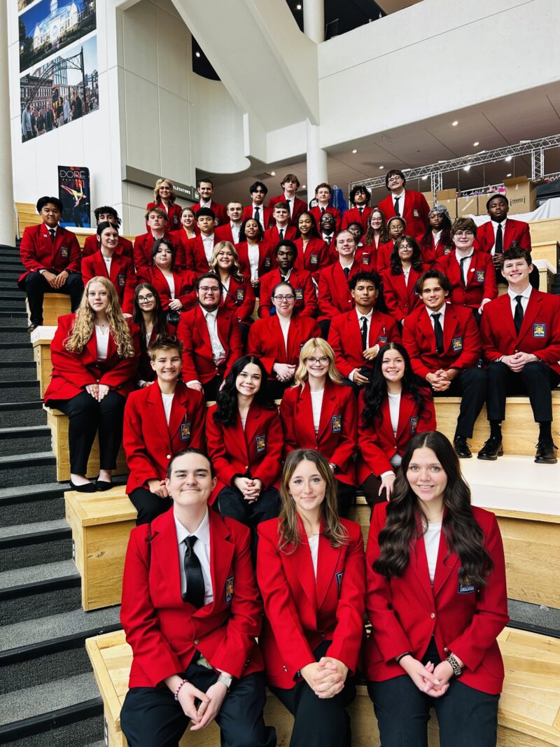 A group of SkillsUSA students, including a regional officer, wearing matching red blazers pose together on wooden bleachers inside a modern building with white walls, large windows, and banners in the background.