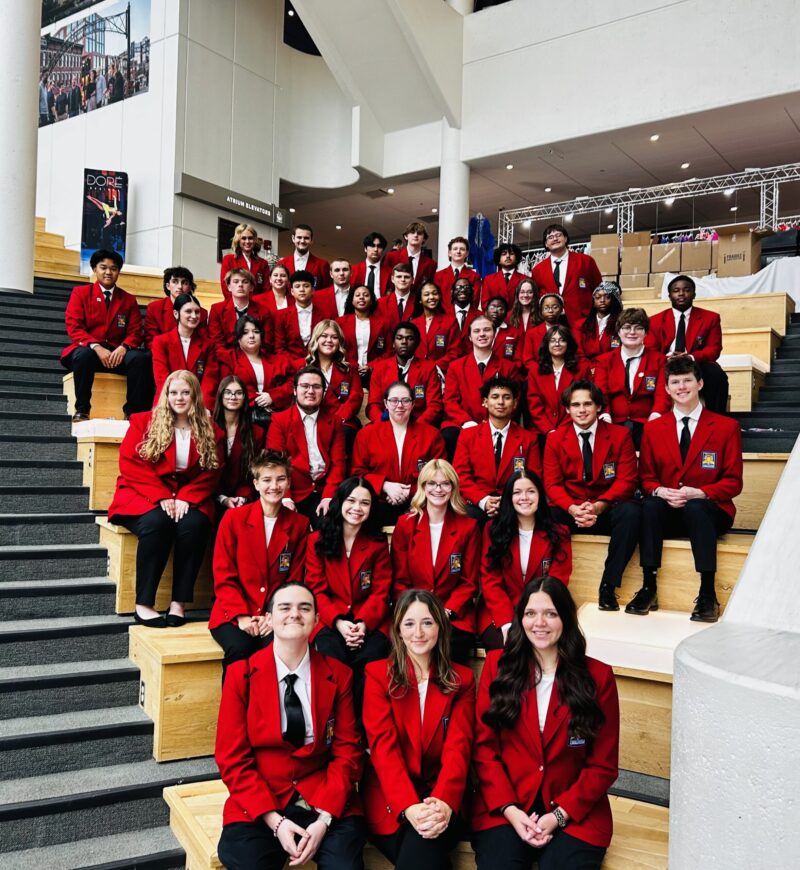 A large group of students, all SkillsUSA Regional Officer candidates in matching red blazers, pose together—seated and standing on wooden steps inside a modern BT building with white walls and large windows.