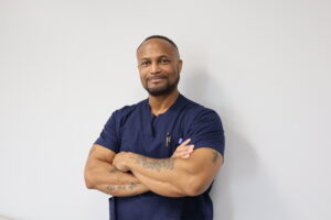 Alumni Spotlight: Kenneth Hall, a Licensed Practical Nursing graduate, stands against a white wall in navy blue scrubs. With short hair, a trimmed beard, visible tattoos, and a pen clipped to his pocket, he smiles confidently.