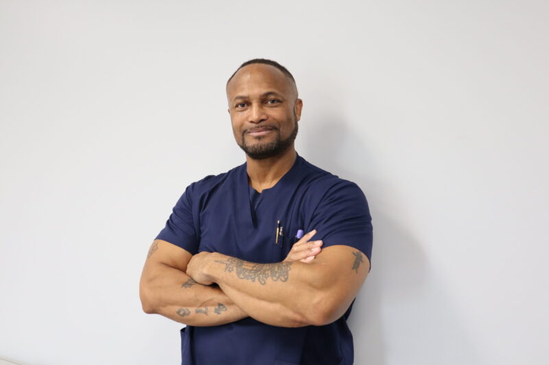 Alumni Spotlight: Kenneth Hall, a Licensed Practical Nursing graduate, stands against a white wall in navy blue scrubs. With short hair, a trimmed beard, visible tattoos, and a pen clipped to his pocket, he smiles confidently.