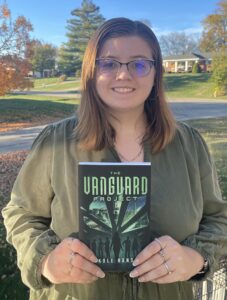 A woman with glasses and shoulder-length brown hair stands outdoors, smiling and holding a book titled "The Vanguard Project" by Kole Hanson. Trees with autumn leaves and a house are visible in the background.