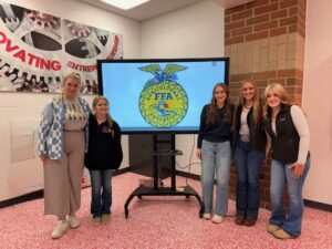 Five smiling young women stand in front of a monitor displaying the FFA logo in a classroom with a pink-patterned floor, showcasing their Madison FFA chapter's dedication to student leadership and decorated, welcoming walls.