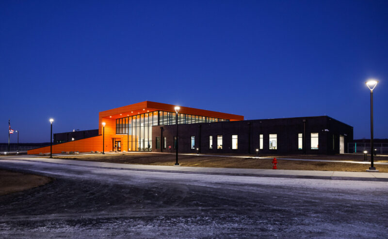 A modern Aviation Center with an orange entrance and large windows is illuminated at dusk, surrounded by streetlights. A flagpole and a fire hydrant are visible outside on the paved area.