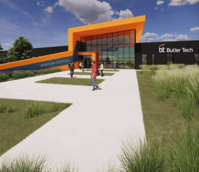 A modern Aviation Center labeled Butler Tech features a bold orange entrance. Several people walk on the sidewalk leading to the entrance, surrounded by grass and landscaping under a partly cloudy sky.