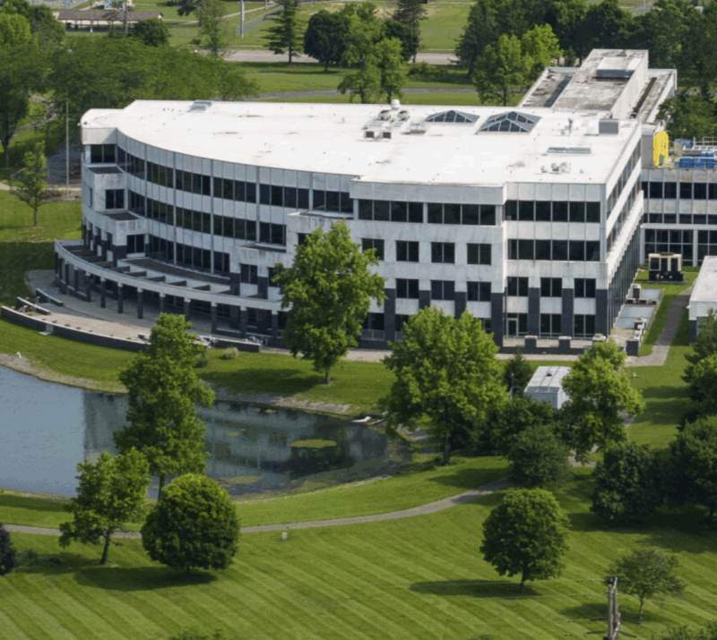 Aerial view of a modern, multi-story office building at the heart of an advanced manufacturing hub, featuring a white roof, green grass, trees, and a small pond in front.