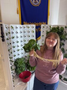 A young person with long hair, representing the Madison FFA chapter, holds up a fresh lettuce plant with roots next to a hydroponic system. A blue and yellow banner hangs in the background, and a red bowl sits on the growing unit.
