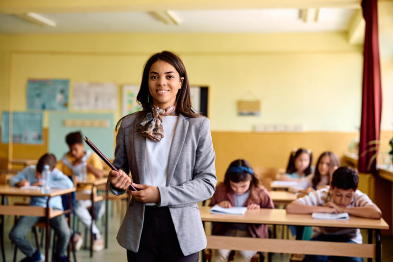 A smiling teacher stands at the front of a bright classroom, holding a tablet, while young students focus on their work. This engaging scene highlights the success of our Teacher Pathway program in real classroom settings.