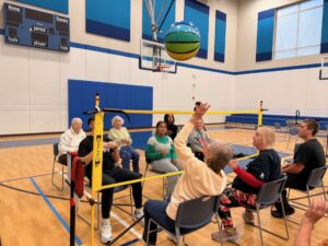 A group of older adults sit in chairs in a gym, playing a seated volleyball-style game with a ball and net. They appear engaged and active, building bonds across generations, with a basketball hoop and scoreboard in the background.