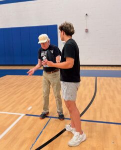 An older man and a younger man stand and talk on a gymnasium basketball court, reflecting strong student bonds across generations. The older man wears a cap and dark shirt; the younger, a black T-shirt and shorts. Blue wall padding lines the background.