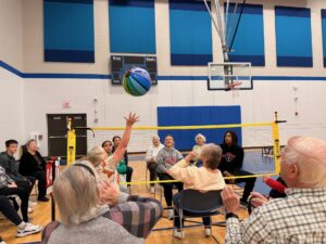 A group of seniors sits in a circle playing seated volleyball with a colorful ball in a gym. A basketball hoop is in the background, as younger people watch from the side, highlighting bonds between generations.
