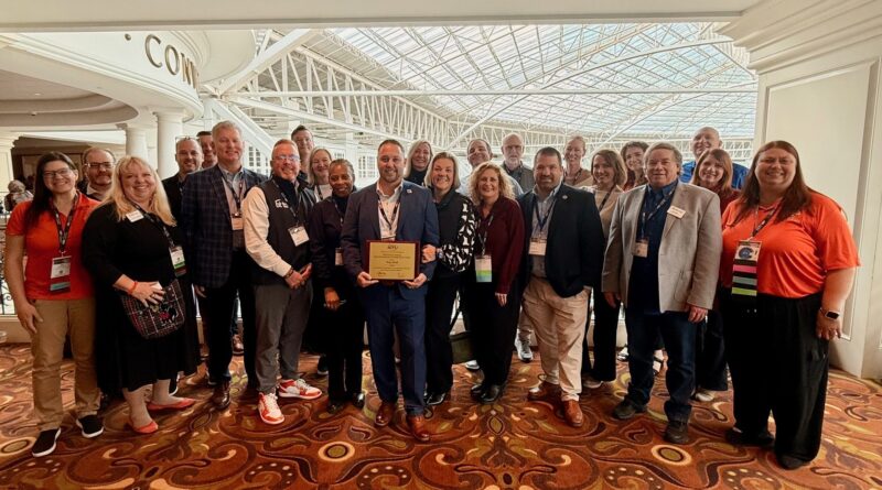 A large group of adults, both men and women, stand indoors posing for a photo. One person in the center holds the National CTE 