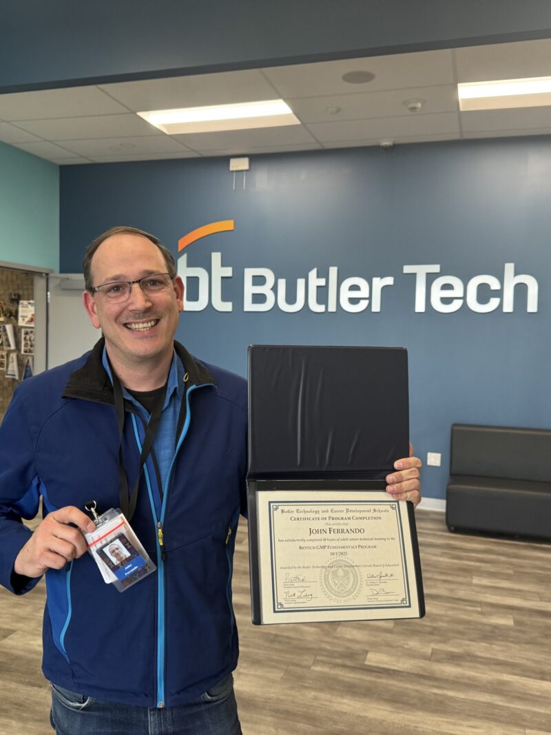 A smiling man stands indoors holding an open certificate and his ID badge, celebrating his achievements in the cGMP cohort. Behind him is a blue wall with the 