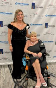 Two women in elegant black gowns—one standing, one seated in a wheelchair—smile together at the 2025 Women of Excellence event, held by Butler Tech, in front of a backdrop featuring the Chamber Alliance and Women of Excellence logos.