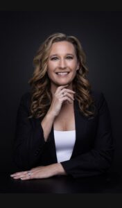 A 2025 Women of Excellence honoree with long curly hair, wearing a black blazer and white top, sits confidently against a dark background—smiling as she represents Butler Tech with one hand under her chin and the other on the table.