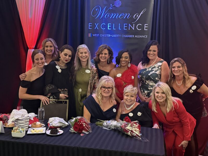 A group of women dressed formally poses and smiles in front of a “Women of Excellence 2025” banner at the Butler Tech event; some hold bouquets, and a table with flowers and food is set in front of them.