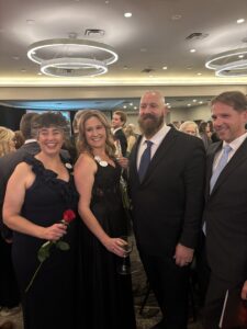 Four people dressed formally at an indoor event, smiling at the camera. Celebrating Women of Excellence 2025 with Butler Tech, one woman holds a red rose and another a wine glass. Other guests and modern ceiling lights fill the background.