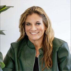 A woman with long, wavy blonde hair is smiling. She is wearing a dark green jacket over a black top and large hoop earrings, representing Women of Excellence at Butler Tech 2025. She sits in front of a white background with a leafy plant to the side.