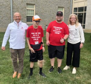 Four people stand smiling on grass in front of a stone building. Two wear matching red "Volunteer Assistance Crew" shirts, representing Miami University’s commitment to Inclusion and programs like Project SEARCH.
