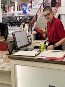 A young man in a red shirt stands behind a front desk with a computer, clipboard, and water bottle. The setting appears to be a gym or fitness center at Miami University, with exercise equipment and banners visible in the background.