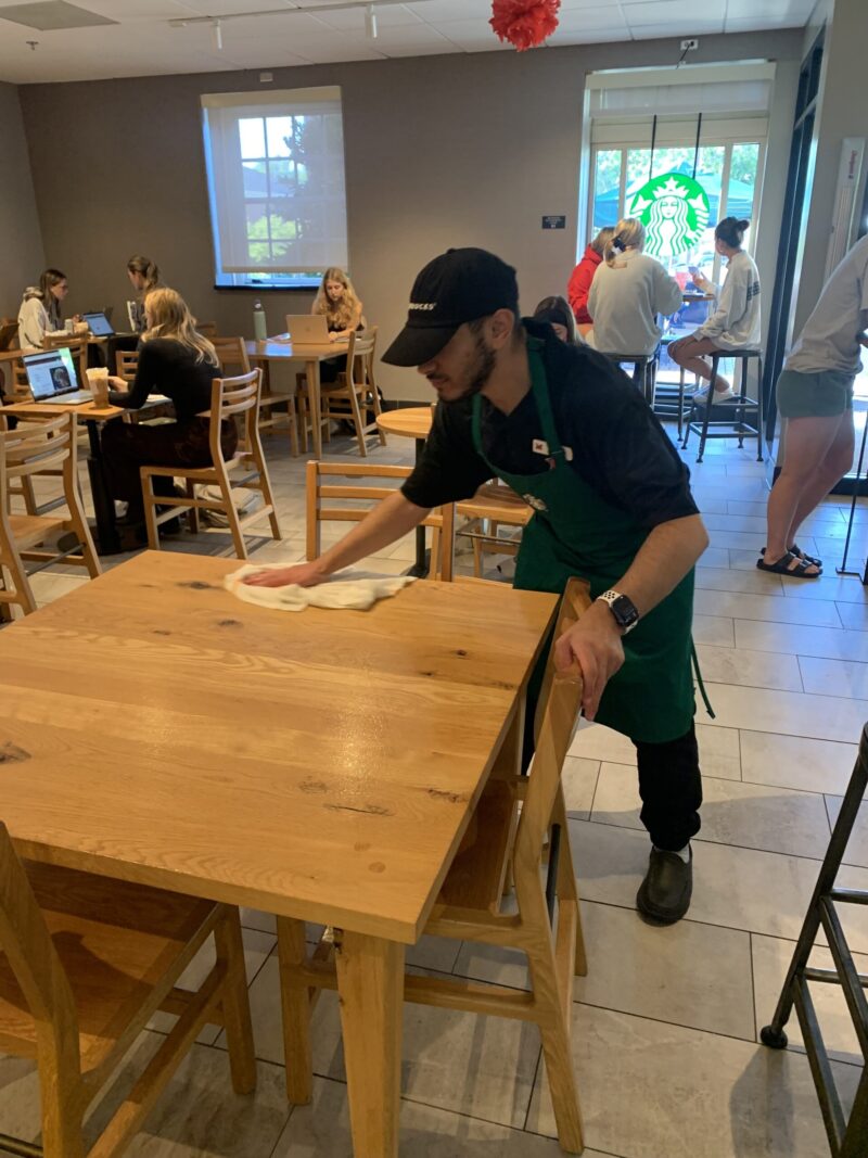 A barista wearing a black hat and green apron wipes down a wooden table in a busy Starbucks café at Miami University, fostering inclusion as customers sit and work in the background.