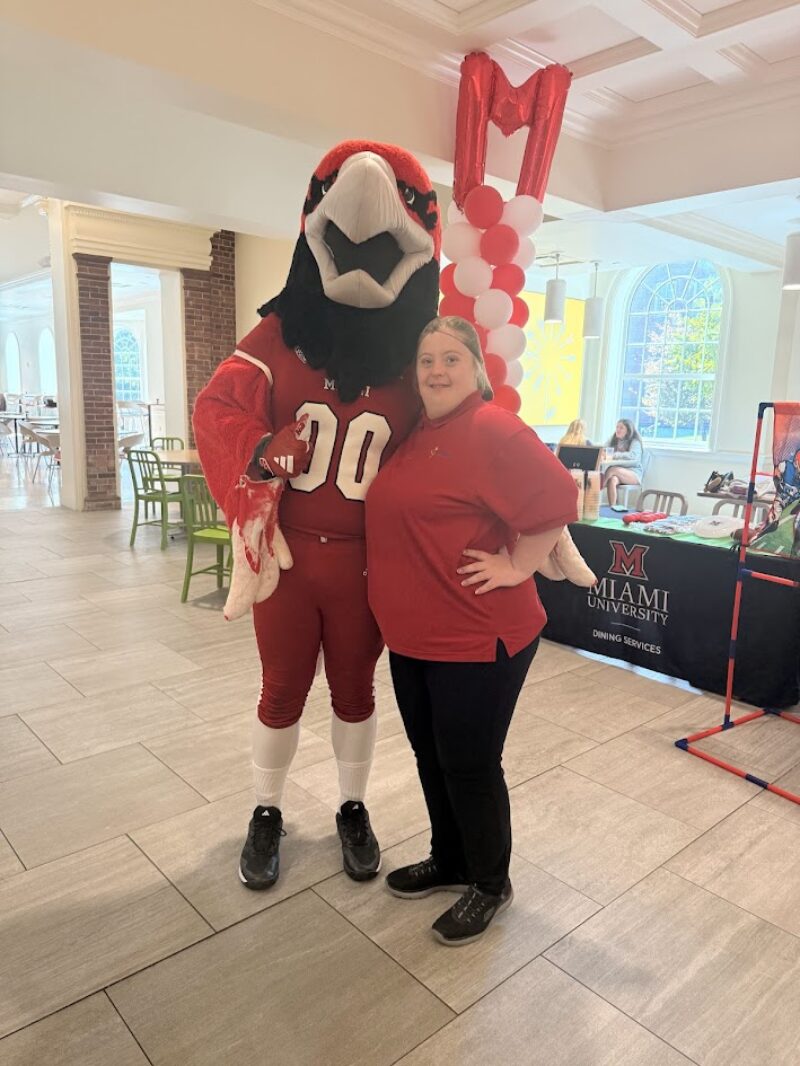 A woman in a red shirt poses next to a person in a red hawk mascot costume inside a bright room with large windows. Behind them are a balloon arch and a Miami University Dining Services table, celebrating inclusion and Project SEARCH.