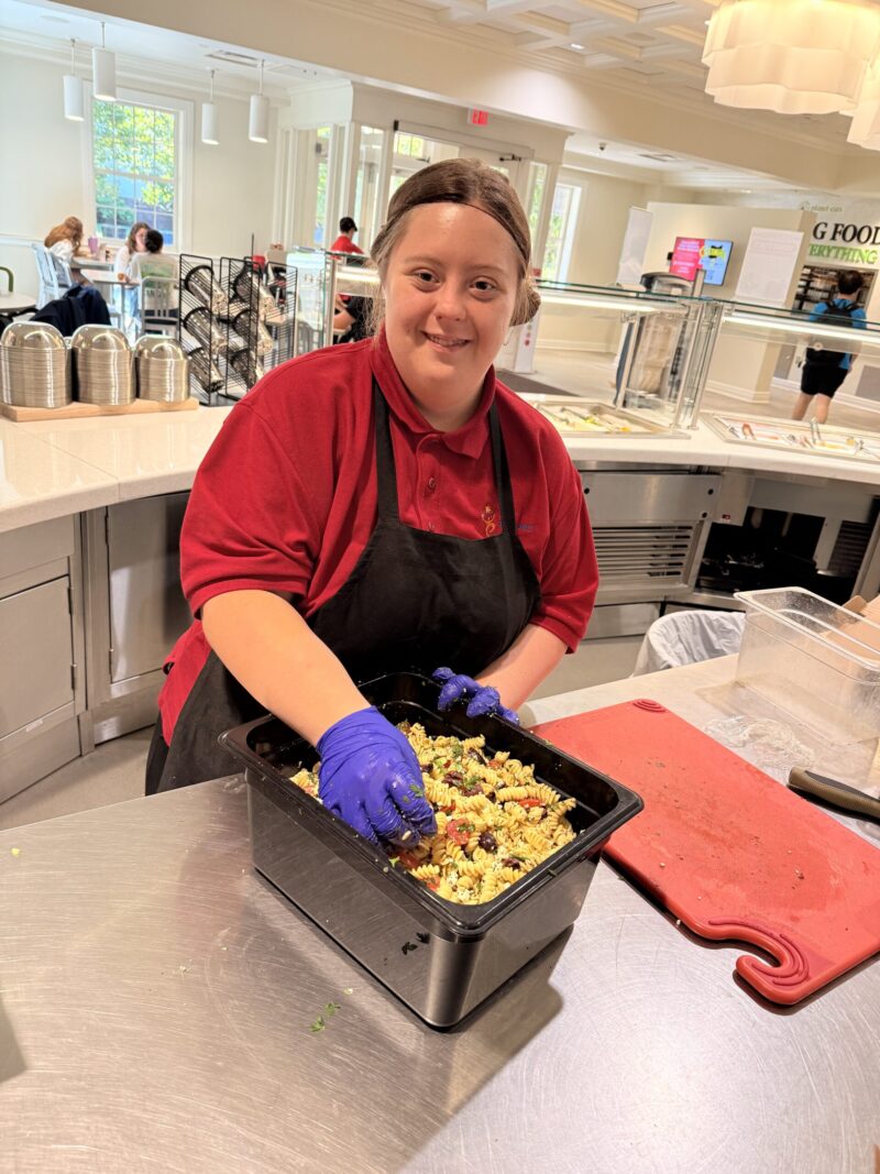 A smiling woman wearing a red shirt, black apron, and blue gloves prepares food in a cafeteria kitchen as part of Project SEARCH, mixing ingredients in a black container. A bright, modern dining area is visible in the background.