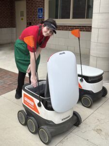 A person wearing a green apron and black visor bends down to load a Grubhub food delivery robot outside a building at Miami University, highlighting efforts toward greater inclusion. Another identical delivery robot is nearby.