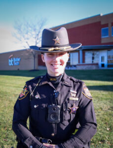 A smiling sheriff’s deputy, Deputy Kinlee Hoyle, stands outside on grass in uniform with a hat and body camera. A building with “Butler Tech” signage is visible in the background. Alumni Spotlight highlights her Basic Police Academy journey.