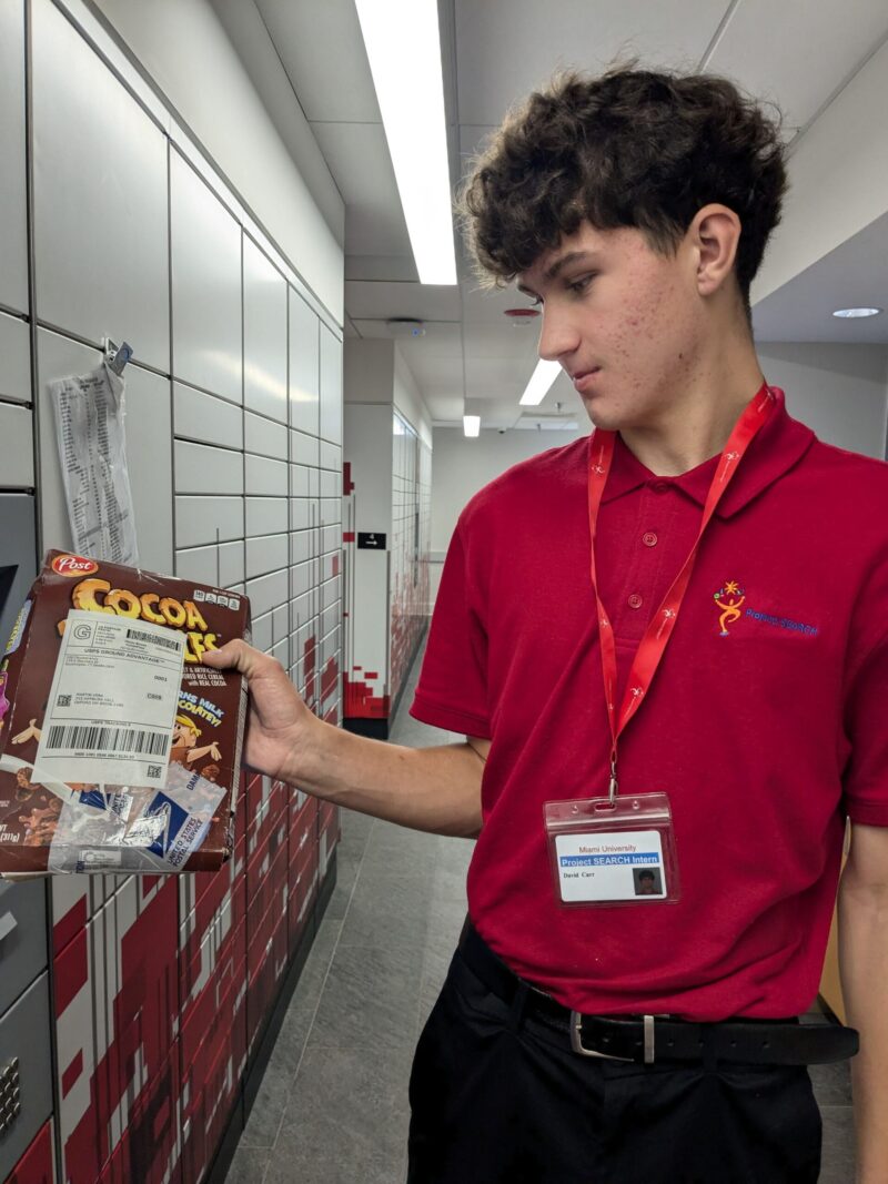 A young man in a red polo shirt with a name badge examines a package containing a Cocoa Pebbles cereal box and mail in front of lockers at Miami University, highlighting inclusion in a well-lit hallway.