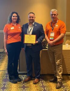 Three people stand indoors. Tony Huff, in a suit, holds a framed National CTE Award certificate. The two on either side, wearing orange CTE for All shirts with badges and logos, face the camera and smile.