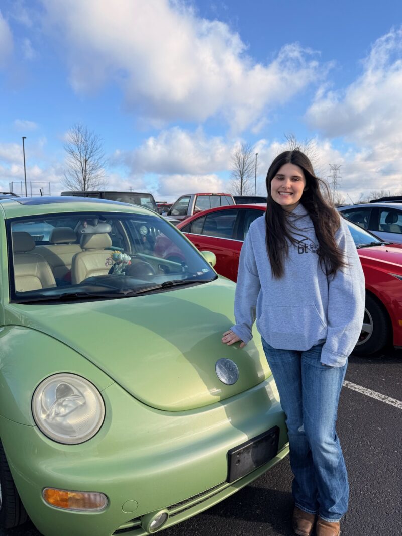 A young woman with long dark hair, wearing a gray hoodie and jeans, stands smiling next to a light green Volkswagen Beetle in a parking lot—a proud moment for student drivers made possible by a grant removing barriers to learning.