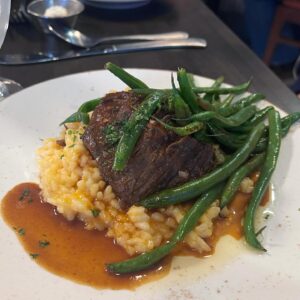 A plate of braised beef served over creamy risotto, topped with sautéed green beans and garnished with herbs, is a savory highlight this season at Café Lee, finished with a rich brown sauce drizzled around the dish.